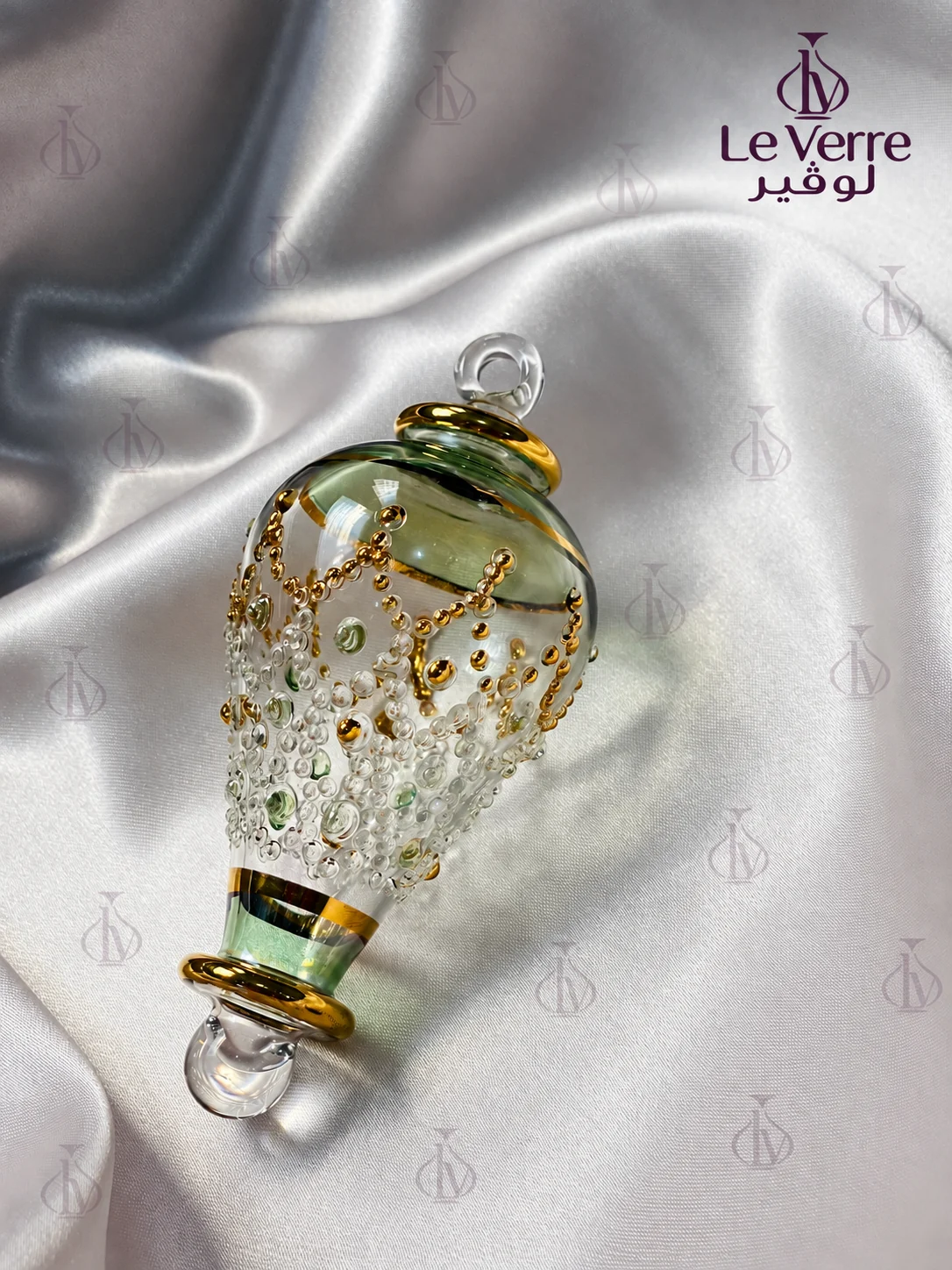A single tapered glass Christmas ornament in clear and pale green glass with gold-tone bands and raised gold-tone and green dot detail, with a clear hanging loop, on white silk