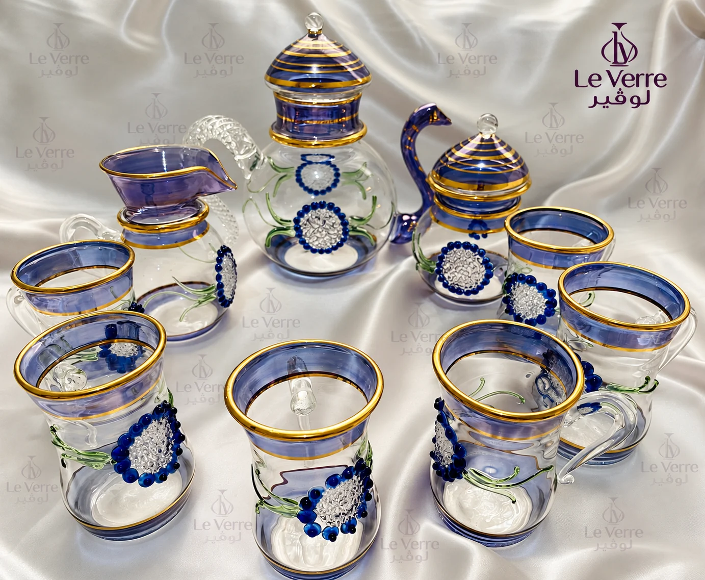 Blue-tinted glass tea and serving set with gold-tone rims and raised blue floral roundels on green painted stems: a teapot, a lidded pot, a creamer jug and six tea glasses on white silk
